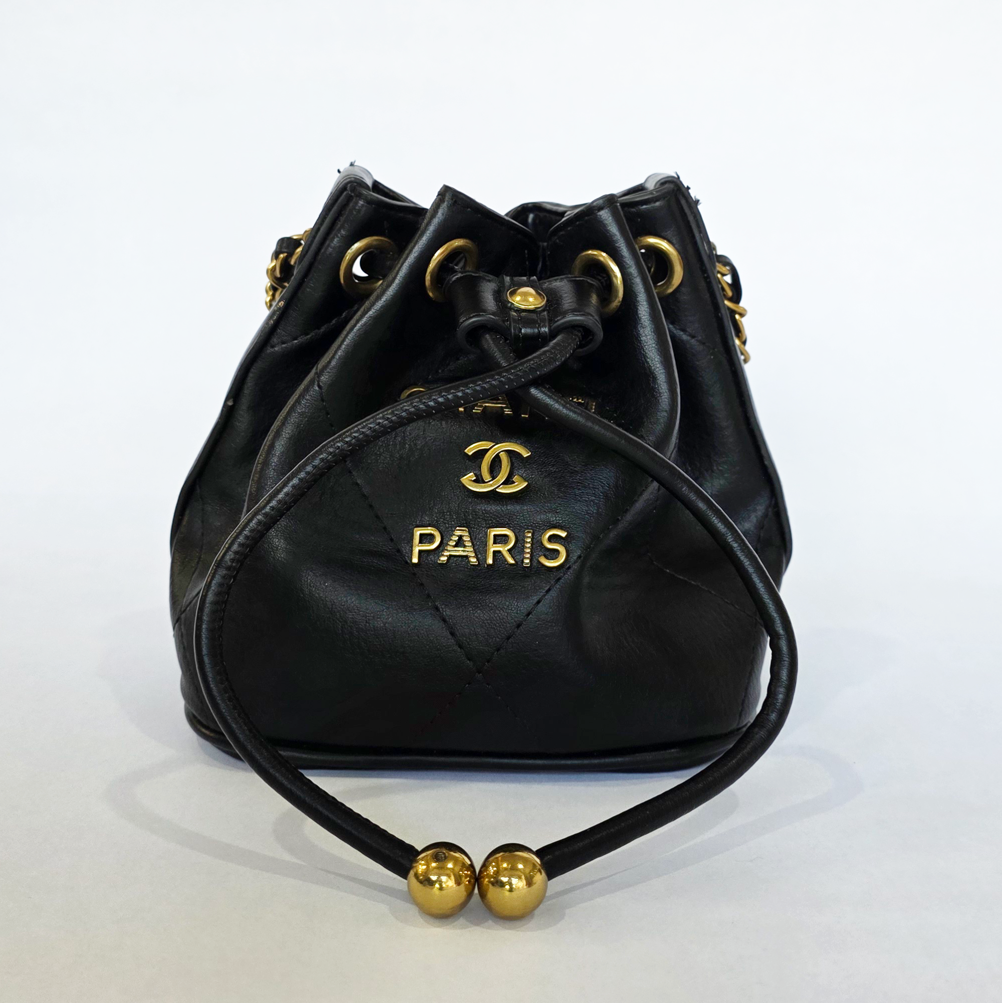 pre loved designer inspired black quilted bucket bag Pre-loved black bucket bag featuring a quilted lower panel, gold lettering, and a drawstring closure with gold balls.